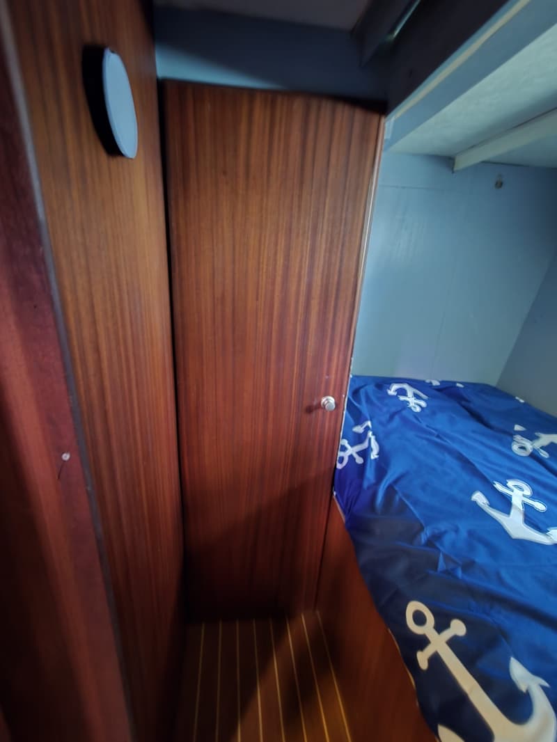 Een kajuit met houten wanden en vloer, een blauw bed met ankerpatroon en een witte ronde lamp aan de muur.
