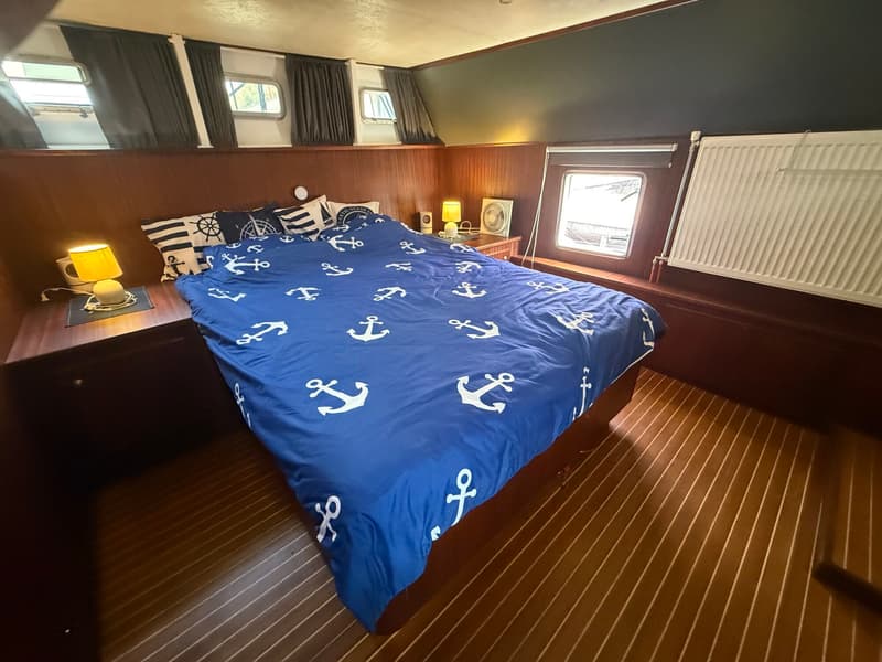 Een slaapkamer met een tweepersoonsbed met blauw beddengoed met ankerpatroon, houten wanden en vloer, en ramen met gordijnen.