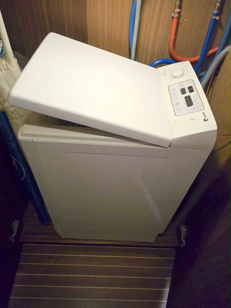 Een witte bovenlader wasmachine met een open deksel staat op een houten vloer met leidingen op de achtergrond.