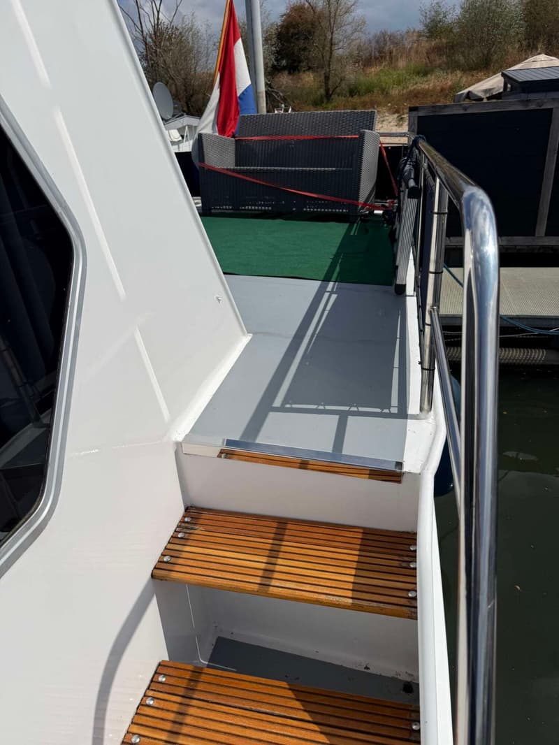 Een witte boot met houten treden en een metalen reling, met een Nederlandse vlag op de achtergrond.