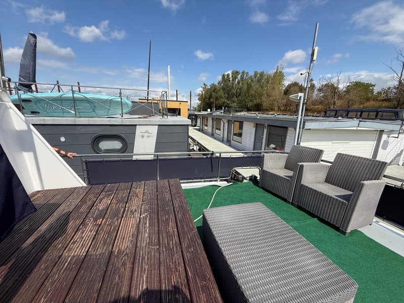 Een bovenaanzicht van een dek met houten planken, groen tapijt, rieten meubels en een supboard op een boot.