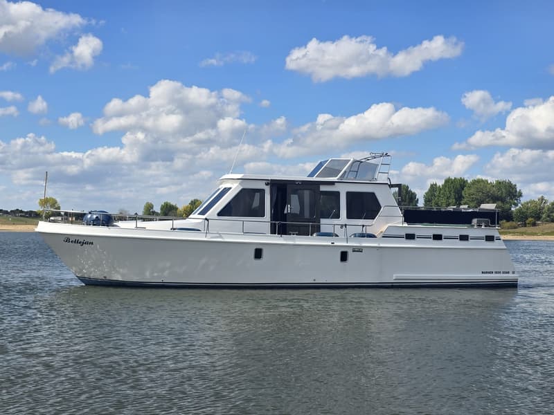Een witte motorboot met de naam "Bellejan" op de boeg en "MARHEN 1500 GSAK" op de achtersteven, vaart op water onder een…