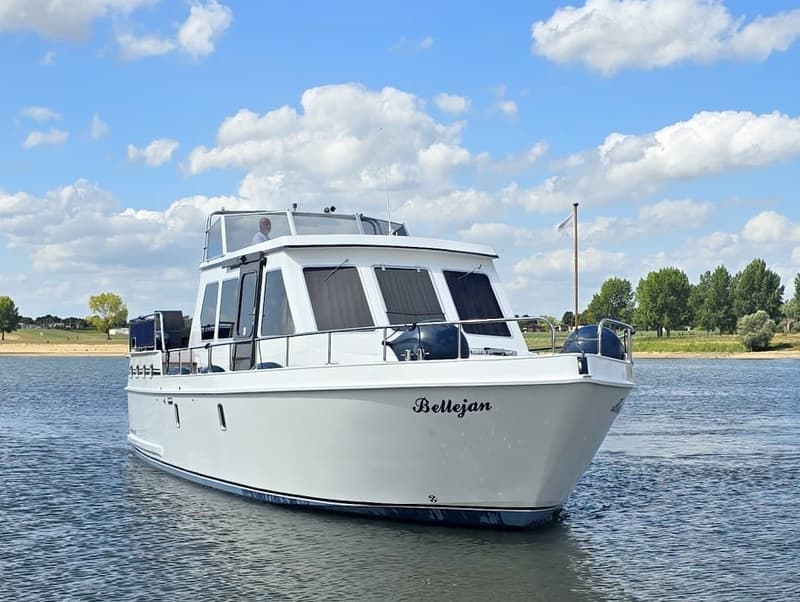 Een witte motorboot met de naam "Bellejan" vaart op het water onder een blauwe hemel met wolken.