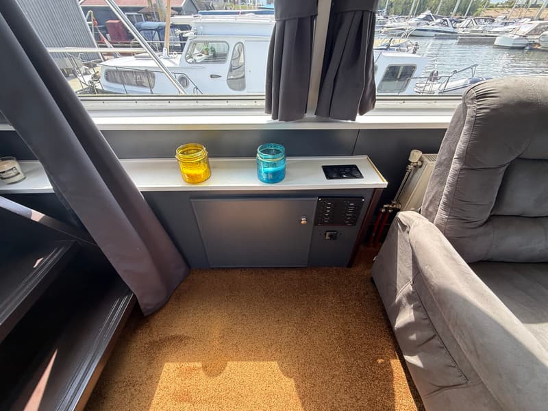 Een interieurfoto van een boot met uitzicht op een jachthaven, met een grijze fauteuil, bruin tapijt en een bedieningspaneel.