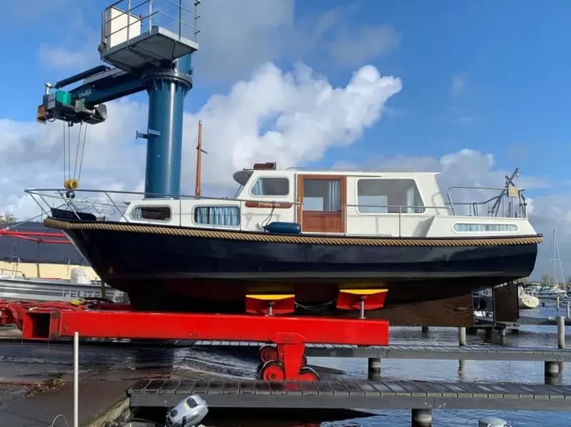 Een donkerblauwe boot met witte opbouw staat op een rode trailer naast een kraan, met een blauwe lucht en wolken.