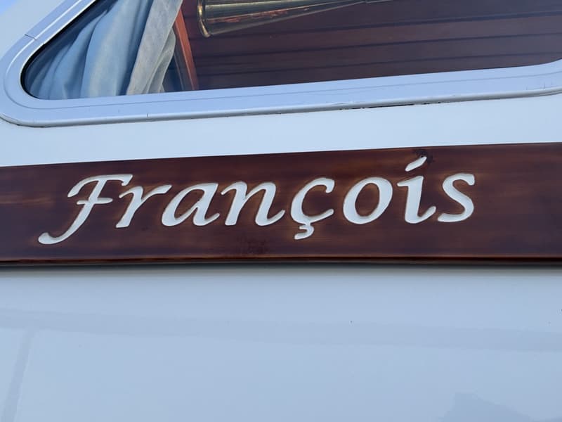 Een witte boot met een bruin houten paneel waarop de naam "François" in witte letters staat, met daarboven een raam.