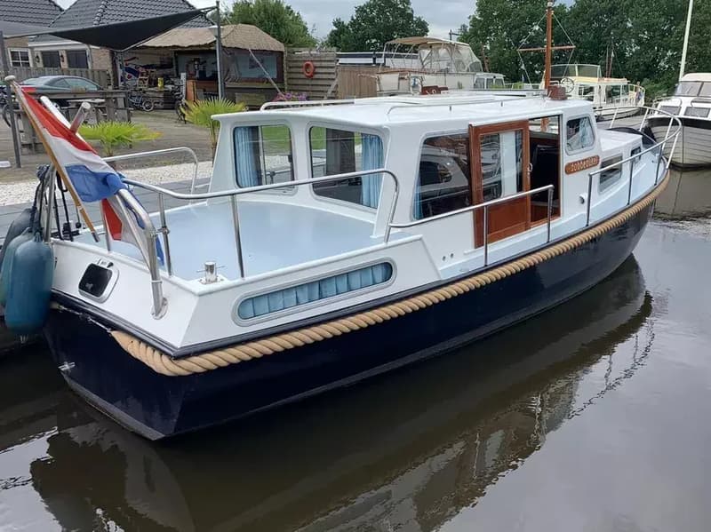 Een witte boot met een donkerblauwe romp en een Nederlandse vlag aan de achtersteven ligt aangemeerd in het water.