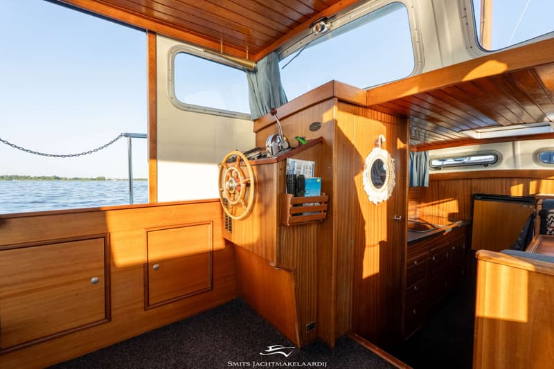 De foto toont het interieur van een boot met een houten stuurwiel, bedieningspaneel en uitzicht op water.