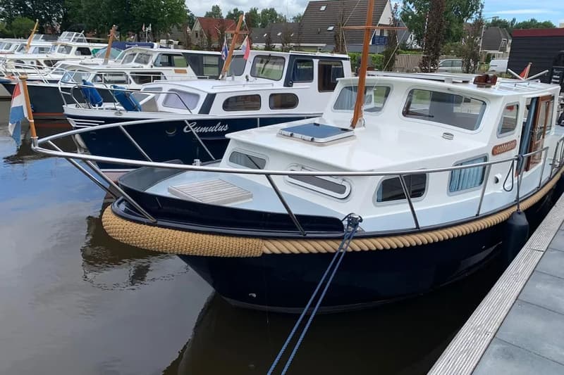 Een witte en donkerblauwe boot met een touwstootrand ligt aangemeerd aan een houten steiger, met andere boten op de…