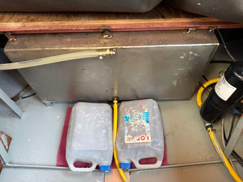 Een metalen tank met slangen, twee plastic jerrycans en een zwarte pomp zijn zichtbaar op een grijze vloer.