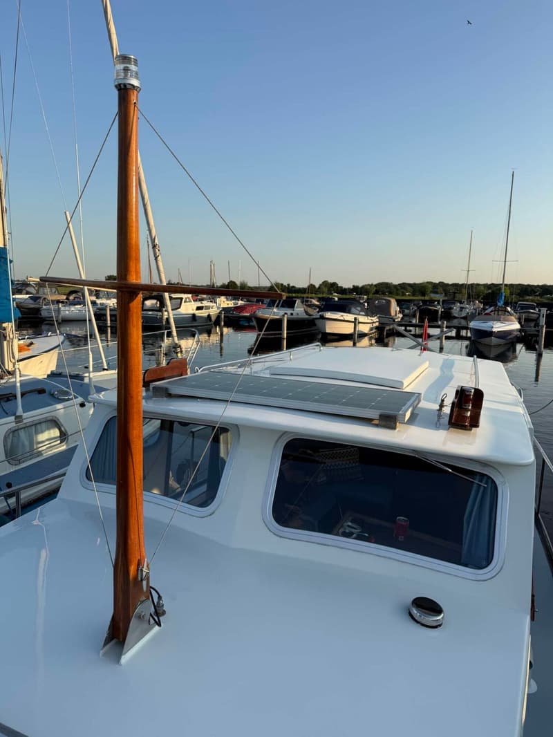 Een witte boot met een houten mast en een zonnepaneel op het dak ligt in een haven vol met andere boten.