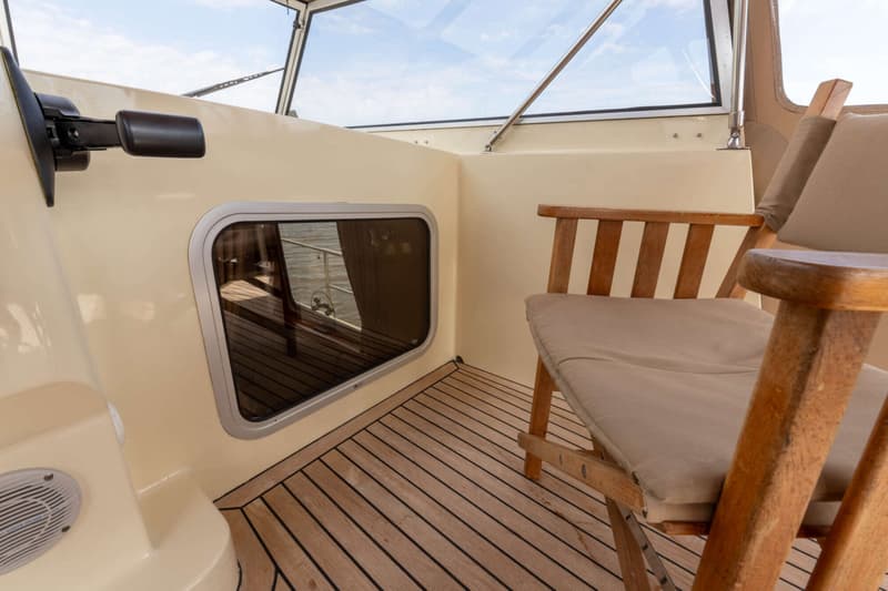 Een houten stoel met beige kussen staat op een teakhouten dek naast een raam en een beige wand.