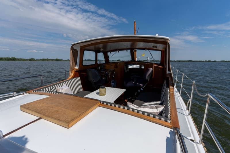 Een boot met een open kuip, houten details en een stuurstand, varend op water onder een blauwe lucht.