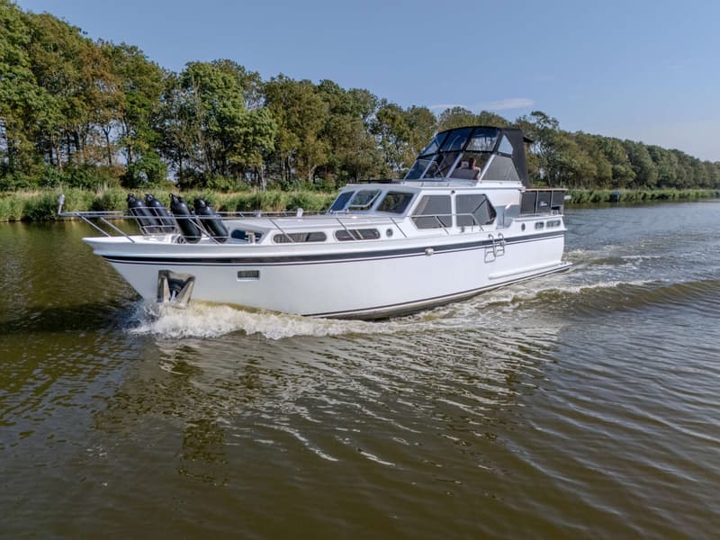 Een witte motorboot met zwarte streep vaart op een waterweg met bomen langs de oever, een persoon is zichtbaar in de…