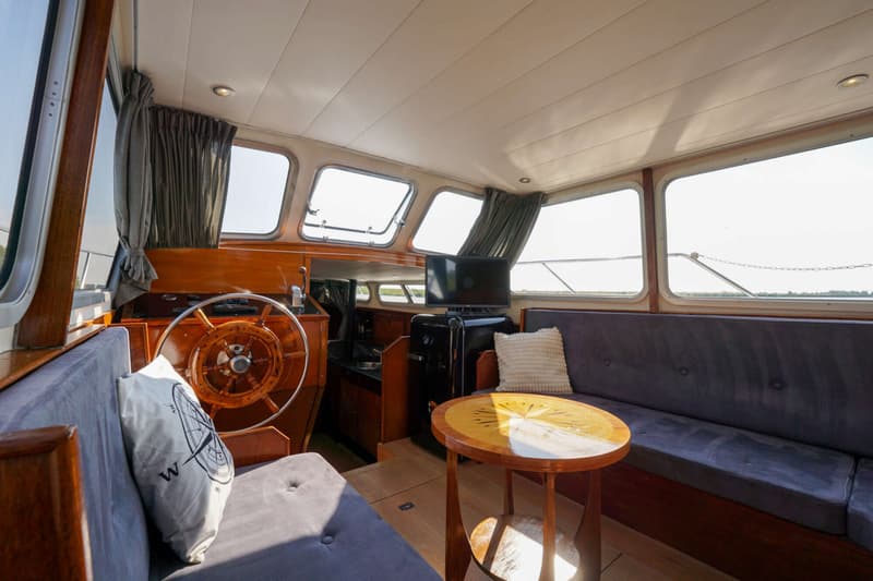 De kajuit van een boot is zichtbaar, met een houten stuurwiel, grijze banken, een tafel met kompasroos en een tv.