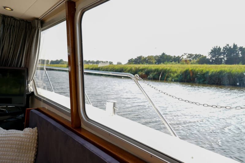 Vanuit een bootraam is een waterweg met groene oevers te zien, met een deel van het interieur en de reling van de boot.