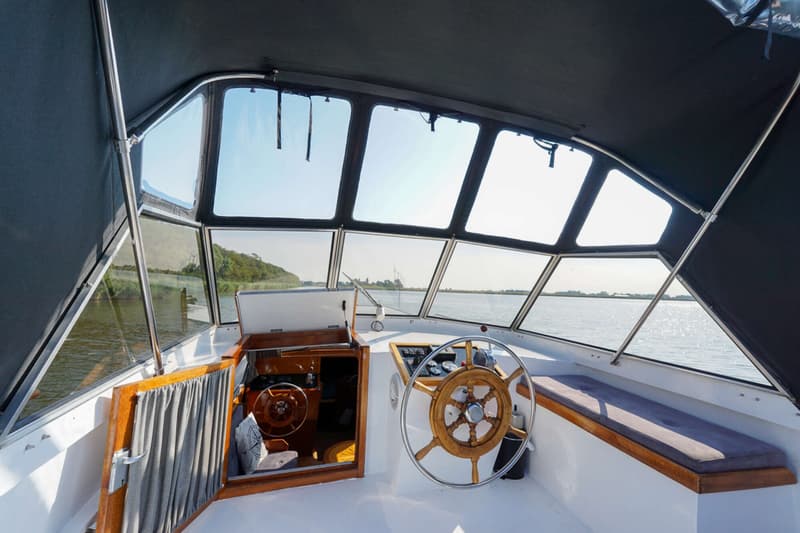 De stuurstand van een boot toont een houten stuurwiel, dashboard, een bankje en de ingang van een kajuit.