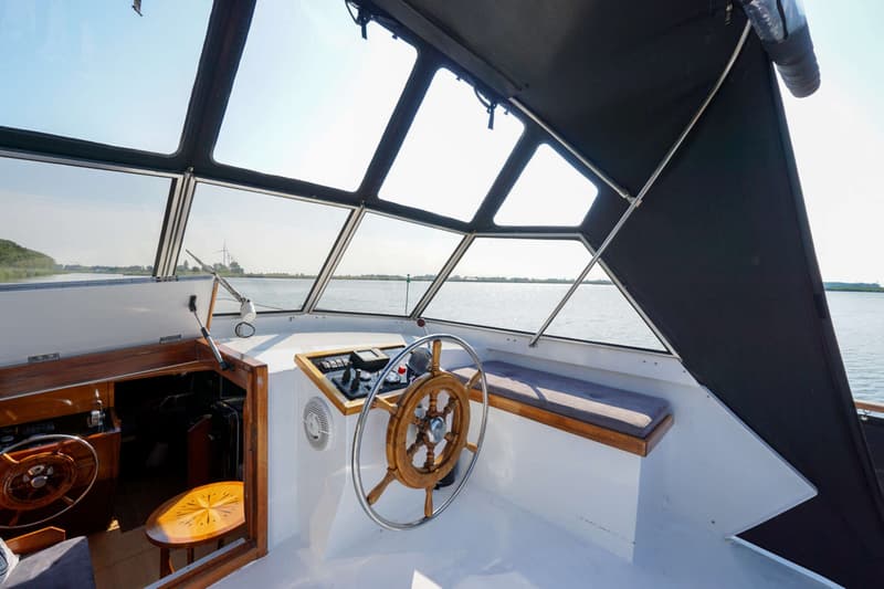 Een houten stuurwiel en dashboard zijn zichtbaar in de open kuip van een boot met grote ramen en een zwarte kap.