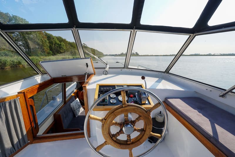 De foto toont de stuurstand van een boot met een houten stuurwiel, instrumentenpaneel en uitzicht over water en landschap.