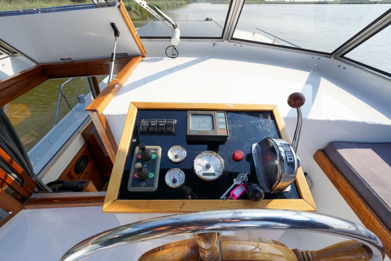 De stuurstand van een boot is zichtbaar, met een houten stuurwiel, bedieningspaneel met meters, schakelaars en een…