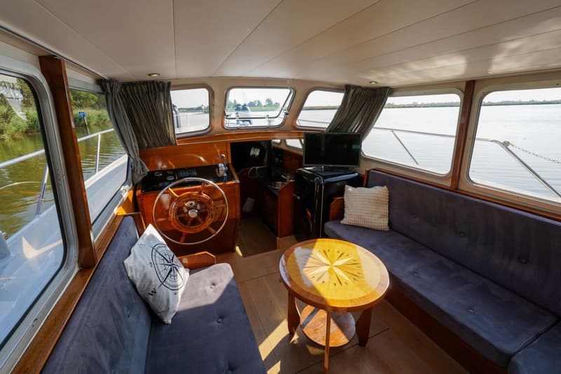 Het interieur van een boot met een houten stuurwiel, twee banken, een ronde tafel, een tv en uitzicht naar buiten.