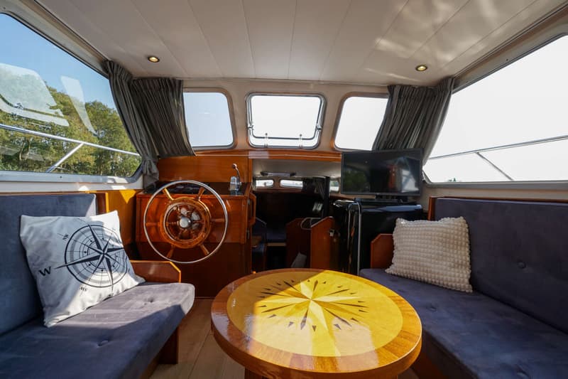 De foto toont een bootinterieur met twee donkergrijze banken, een houten tafel met kompasroos, een stuurwiel en een…