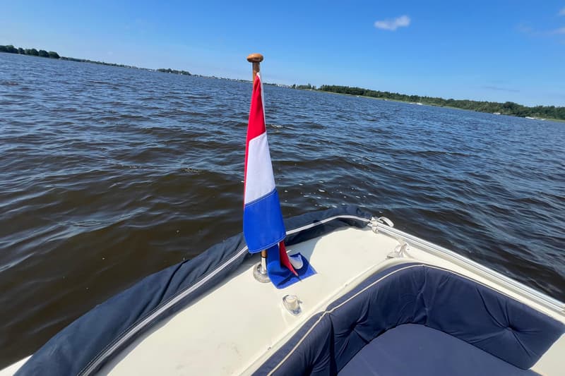 Een Nederlandse vlag wappert aan de achtersteven van een boot op een meer met beboste oevers onder een blauwe lucht.