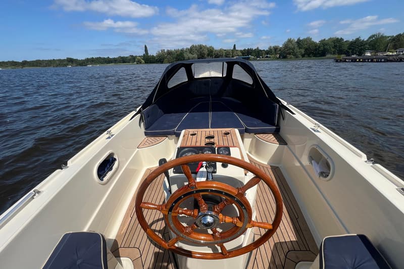 Een boot met een houten stuurwiel en een blauwe kap vaart op een meer met bomen op de achtergrond. — Interboat 19