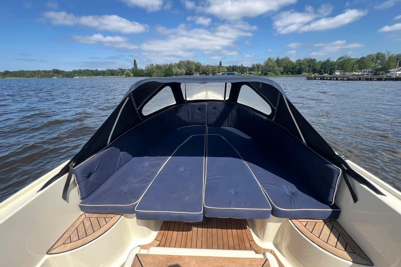 Een boot met een donkerblauwe kap en kussens vaart op een meer met bomen langs de oever onder een blauwe lucht.