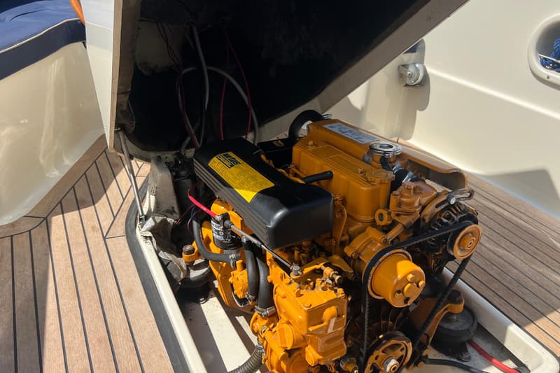 Een gele scheepsmotor met zwarte onderdelen en riemen is zichtbaar, omringd door een houten dek en witte bootonderdelen.