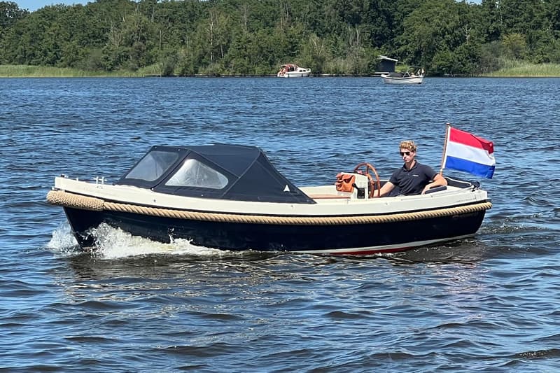 Een donkerblauwe sloep met een crèmekleurige binnenkant en een zwarte kap vaart op het water met een man aan het roer.