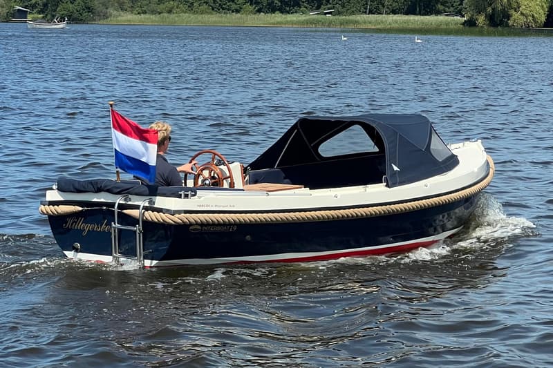 Een donkerblauwe sloep met een witte bovenrand en een rode streep, vaart op het water met een persoon aan het roer.