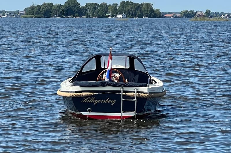 Een donkerblauwe boot met de naam "Hillegersberg" en een Nederlandse vlag, ligt in het water met een zwemtrap aan de…