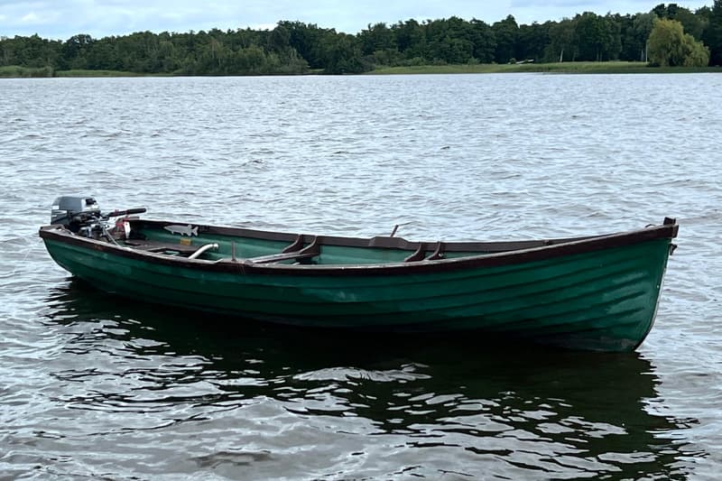 Een groene roeiboot met een buitenboordmotor ligt in het water, met een beboste oever op de achtergrond.