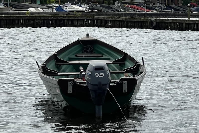 Een donkergroene roeiboot met een Yamaha 9.9 buitenboordmotor ligt in het water, met een steiger en andere boten op de…