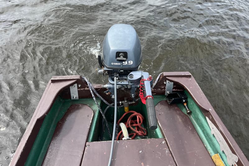Een Yamaha 9.9 buitenboordmotor is gemonteerd op de spiegel van een bruin-groene boot, met water eromheen.
