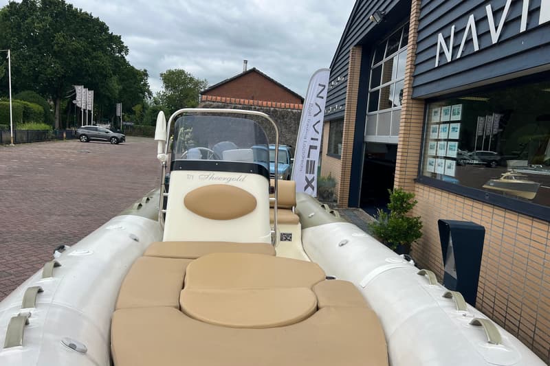 Een witte rubberboot met beige kussens staat voor een gebouw met de tekst "NAVILEX" en een vlag met "Boat Trading…