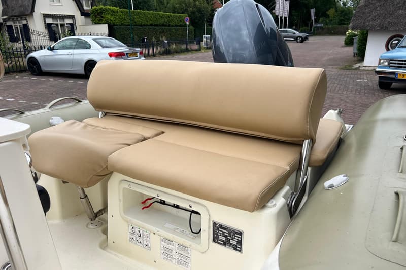 Een boot met beige kussens en een buitenboordmotor, met op de achtergrond een huis en geparkeerde auto's.