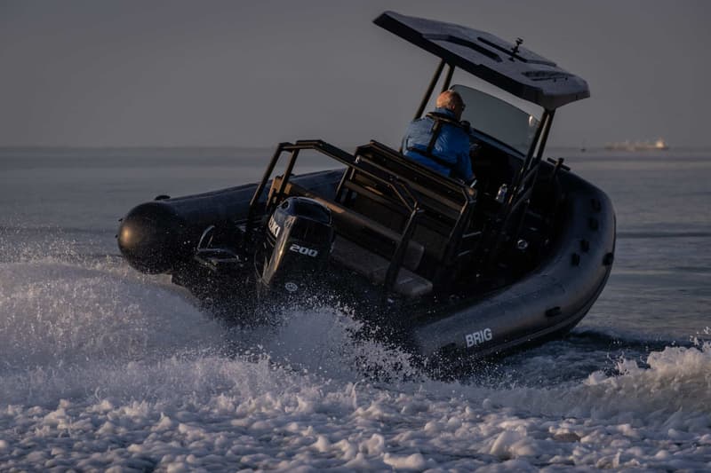 Een man in een blauwe jas bestuurt een zwarte BRIG-RIB met een Suzuki 200 pk buitenboordmotor, die door water vaart.