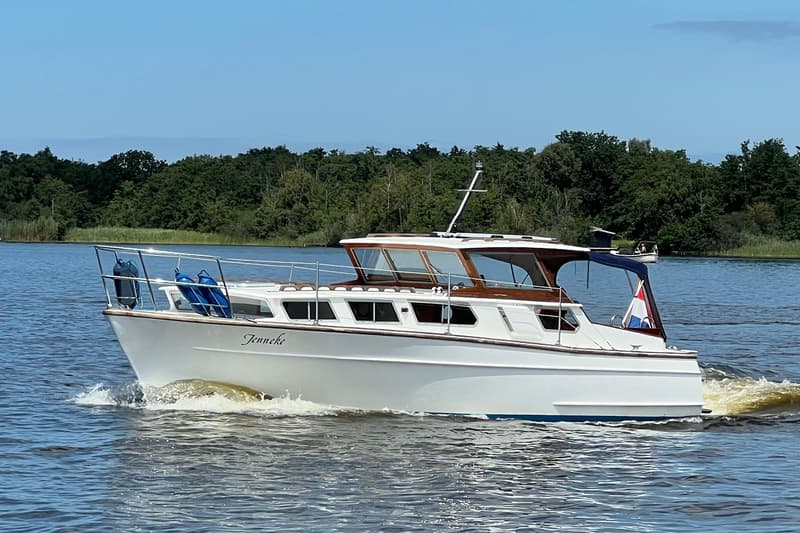 Een witte motorboot met de naam "Jenneke" vaart op het water, met een Nederlandse vlag aan de achterkant.