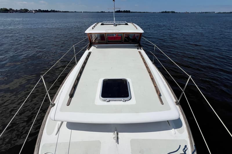 Een bovenaanzicht van een witte boot met een houten stuurhut en een luik op het voordek, varend op rustig water.