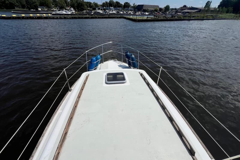 De foto toont de boeg van een boot met twee blauwe stootwillen, een reling en een luik, varend op donker water.
