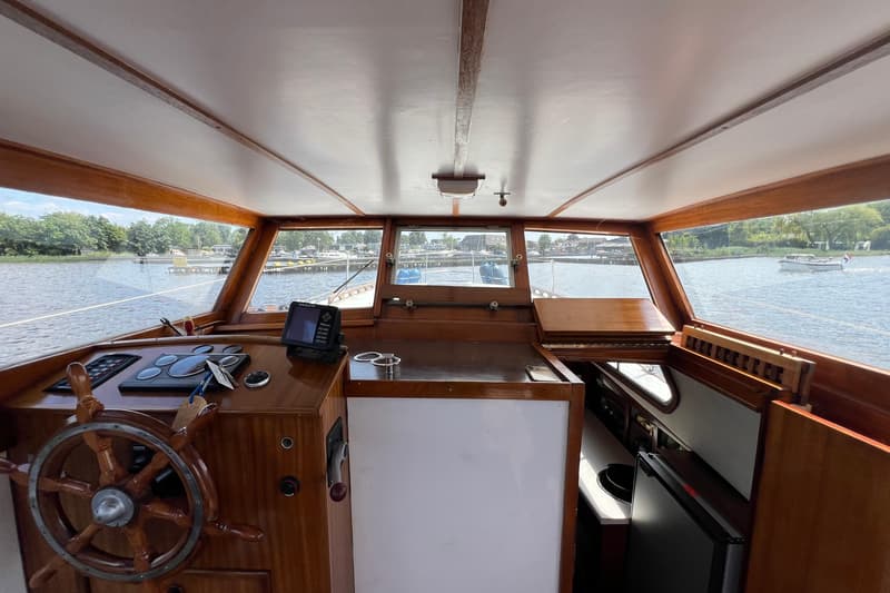 De foto toont het interieur van een boot met een houten stuurwiel, navigatieapparatuur en een uitzicht op het water.