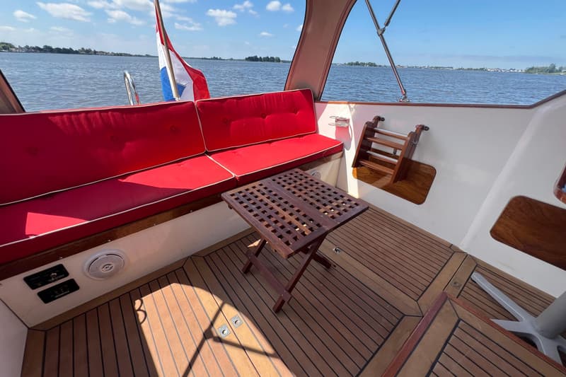 Een bootinterieur met rode kussens, een houten tafel, een Nederlandse vlag en uitzicht op water en landschap.
