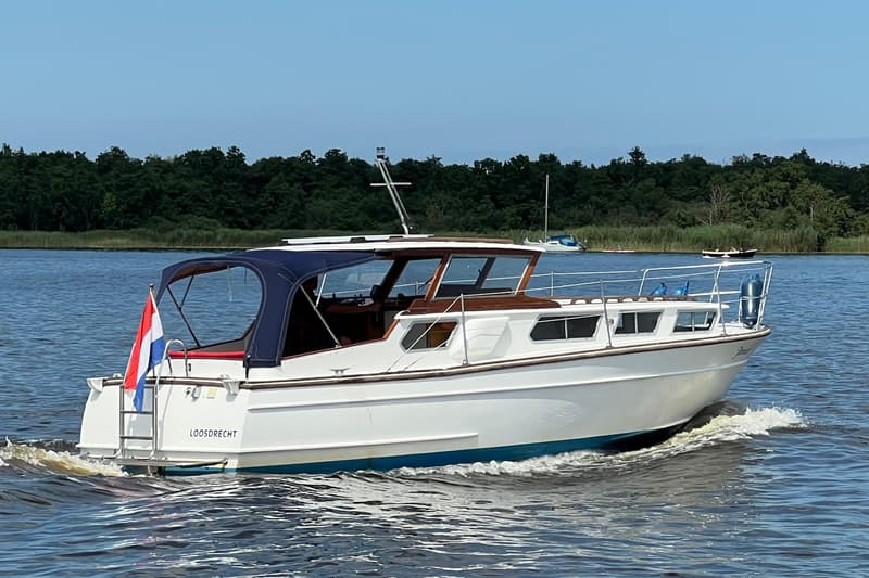 Een witte motorboot met blauwe kap en Nederlandse vlag vaart op het water, met "LOOSDRECHT" op de spiegel.