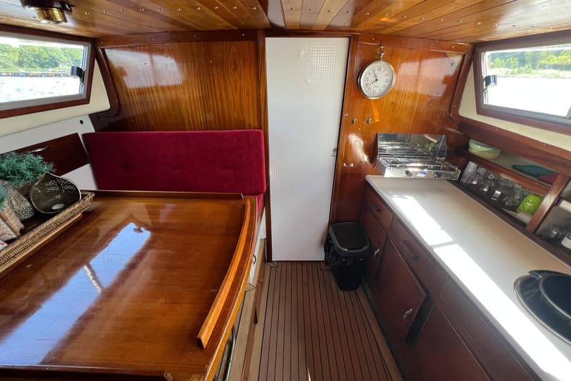 De foto toont de kajuit van een boot met houten wanden en plafond, een rode bank, een tafel, een kombuis en ramen.