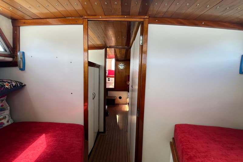 De foto toont het interieur van een boot met houten plafonds en vloeren, witte wanden, rode matrassen en een doorgang.