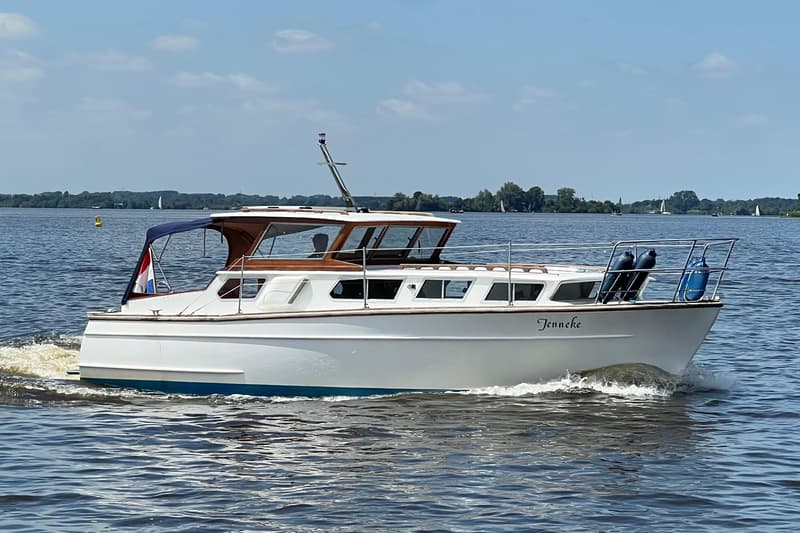 Een witte motorboot met de naam "Jenneke" vaart op een meer met een blauwe hemel en groene oevers op de achtergrond.