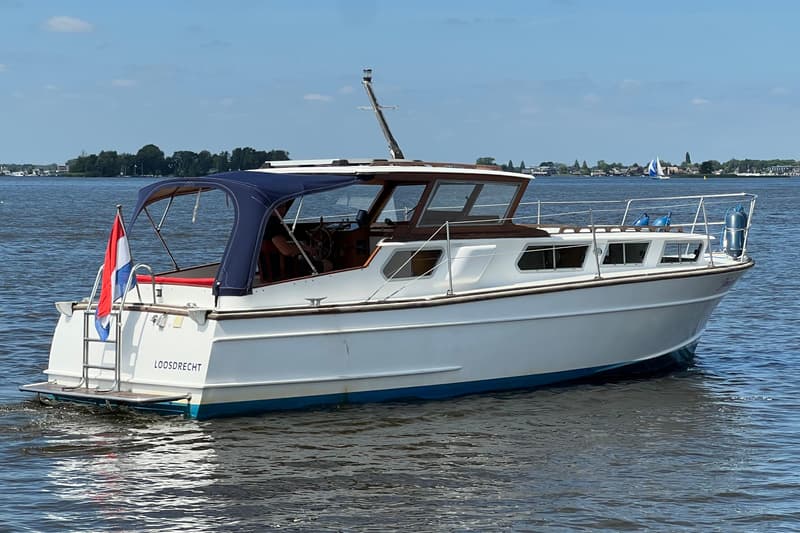 Een witte motorboot met een blauwe kap en een Nederlandse vlag vaart op het water, met "LOOSDRECHT" op de spiegel.