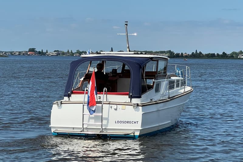 Een witte motorboot met blauwe kap en de naam "LOOSDRECHT" op de spiegel vaart op een waterweg. — Kruiser Polyflash 950